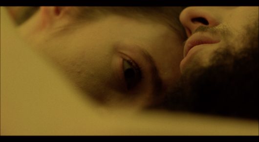 Still from Cambridge Connections: a close view of a man's face seen from the side as he lies down, in warm yellow light.