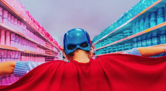 Still from Blow: a person in a blue mask and red cape, seen from behind, faces the aisles of a brightly lit supermarket.