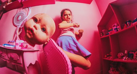 Still from Blow: a girl in a blue skirt lies on a pink floor beside a large doll head, with shelves of toys behind, in bright pink light.