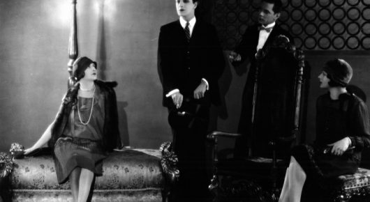 Black-and-white still from The Lodger: women in 1920s dresses and men in dark suits stand and sit in a room with an ornate sofa.