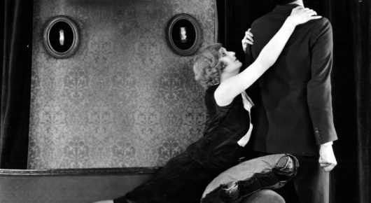 Black-and-white still from The Lodger: a woman in a dark dress leans back in the arms of a man in a dark suit, against a patterned wall.