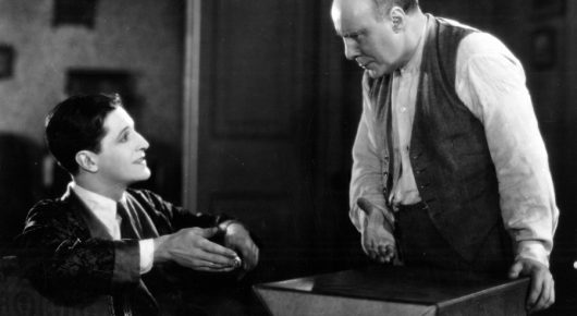 Black-and-white still from The Lodger: a seated man in a dark jacket looks up at an older man in a waistcoat who stands at a table.