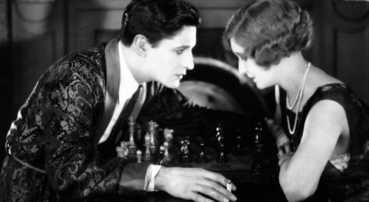 Black-and-white still from The Lodger: a man in a patterned jacket and a woman in a dark dress lean close together over a table with small pieces on it.