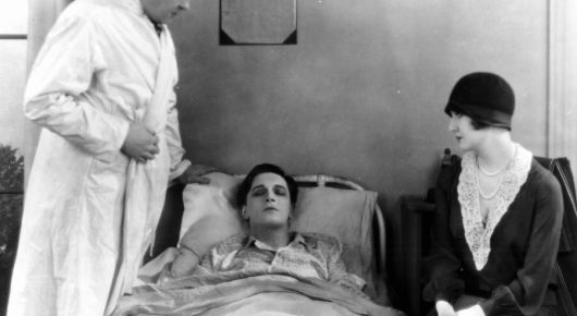 Black-and-white still from The Lodger: a woman lies in bed, with a man in a white nightshirt standing at one side and a woman in a cloche hat sitting at the other.