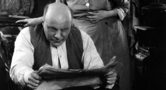 Black-and-white still from The Lodger: a heavyset man in a waistcoat sits reading a newspaper, with a woman standing behind him.