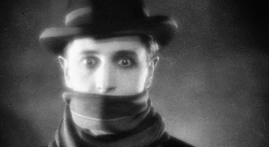 Black-and-white still from The Lodger: a close-up of a man in a hat with a scarf covering the lower half of his face, staring wide-eyed.