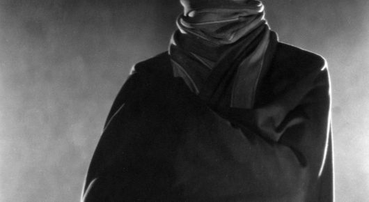 Black-and-white still from The Lodger: a figure in a dark coat and a thick scarf, shown from the chest up with the head out of frame.