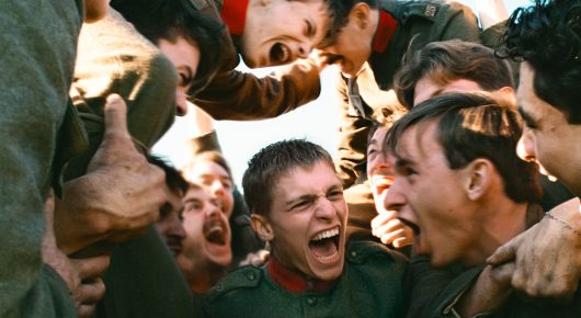 Still from Coward: a young soldier in a green uniform laughs in the middle of a crowd of shouting men in uniform.