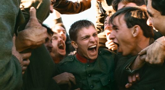 Still from Coward: a young soldier in a green uniform laughs in the middle of a crowd of shouting men in uniform.