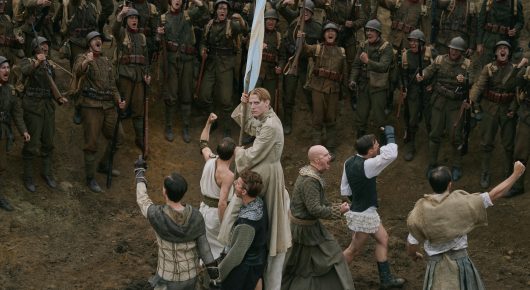 Still from Coward: soldiers in uniform gather in a circle around a muddy clearing where several people, including a figure in a long pale robe, are performing.