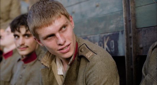 Still from Coward: a young soldier with short fair hair looks up and to one side, with another man in uniform beside him, near a vehicle.