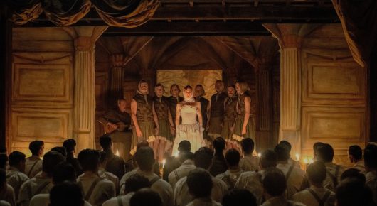 Still from Coward: a crowd of soldiers watches a candlelit stage where a few performers stand in front of ornate columns.