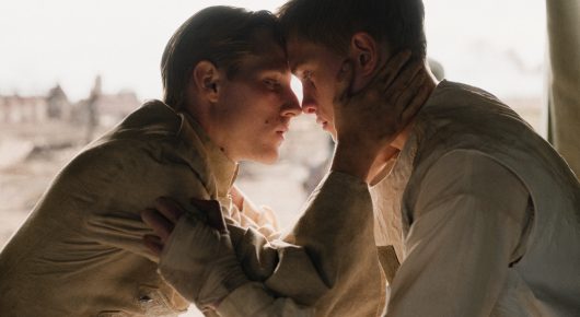Still from Coward: two young men in khaki uniforms press their foreheads together in an embrace against a pale, hazy sky.