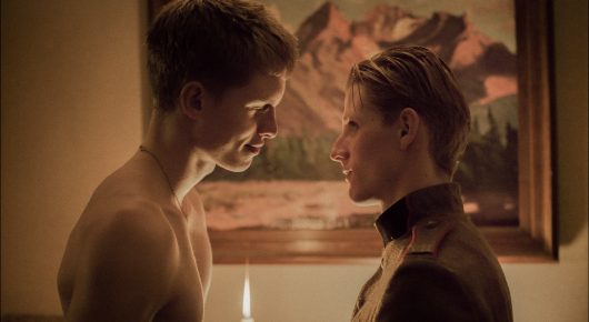 Still from Coward: two young men face each other closely in a warmly lit room, one shirtless and one in a dark uniform, with a framed painting behind them.