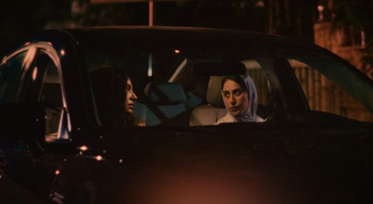 Still from Coyotes: a woman sits in the back of a car at night, seen through the window, with warm light on her face.