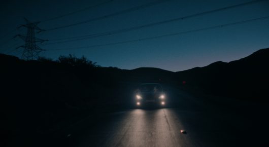 Still from Coyotes: a car with its headlights on drives along a dark road toward the camera, with hills and a pylon against a dusk sky.