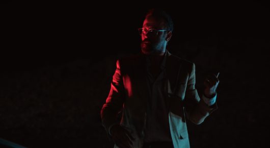 Still from Coyotes: a man stands in darkness, lit by red light from one side.