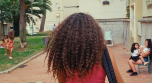 Still from Cura Sana: a girl with long curly hair, seen from behind, walks along a path in a park, with people sitting on a bench in the distance.