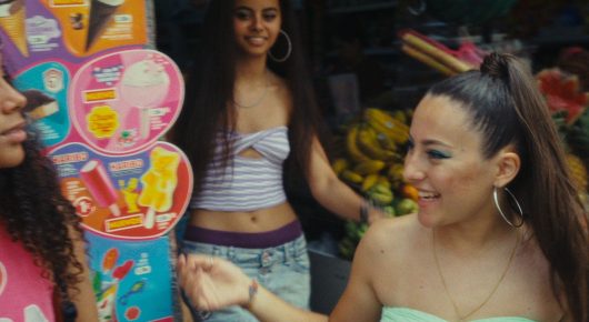 Still from Cura Sana: three young women laugh and talk in front of a colourful shop display of snacks.