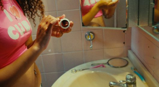 Still from Cura Sana: a woman in a pink top holds a small object above a washbasin in a pink-tiled bathroom.