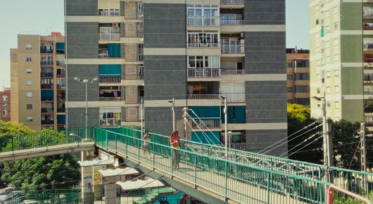Still from Cura Sana: a tall apartment block and a green footbridge over a road, with people in the distance.