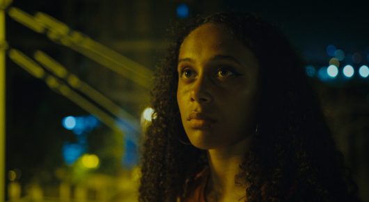 Still from Cura Sana: a woman with long curly hair looks to one side at night, with blurred city lights behind her.