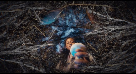 Still from Daughters of the Forest: seen from above, a woman and a child lie among tree roots on the forest floor.