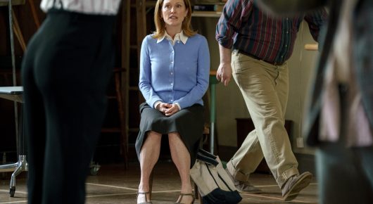 Still from The Debut: a woman in a blue cardigan sits upright on a chair on a stage with her hands in her lap, while people walk past in the foreground.