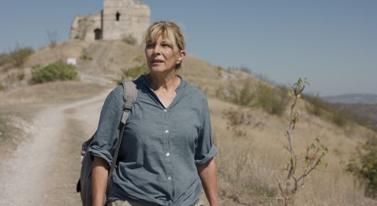 Still from The Dreamed Adventure: a woman in a blue shirt with a rucksack walks along a dry hillside path, with a ruined stone tower behind her.