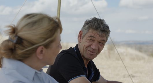Still from The Dreamed Adventure: a woman with blonde hair and a smiling man in a black T-shirt sit close together, with a pole behind them.