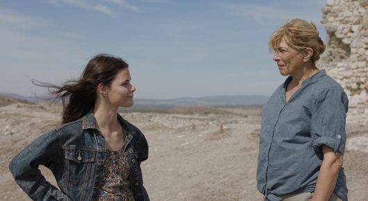 Still from The Dreamed Adventure: a young woman with dark hair in a denim jacket and an older woman in a blue shirt stand facing each other on a dry hilltop.