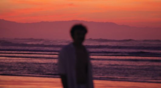 Still from Dado: a blurred figure stands on a beach at dusk under a pink and orange sky, with waves behind.