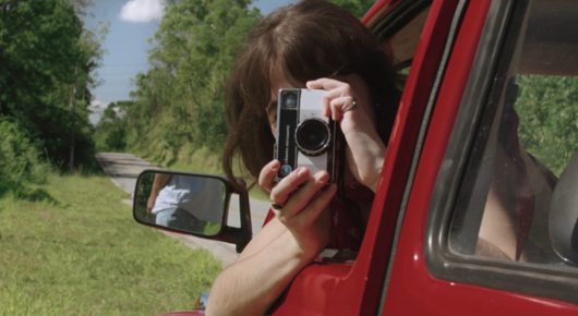 Still from Dado: a woman holds a small camera up to her face from the window of a red car, with green trees behind.