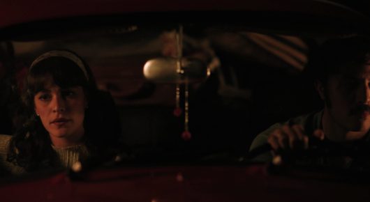 Still from Dado: two people sit in a car at night, with a young woman in a headband in front, lit in red.