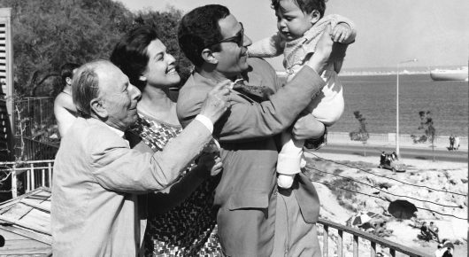 Still from El Verdugo: a black-and-white photograph of a family group outdoors, a man lifting a child while others stand close, with the sea behind them.