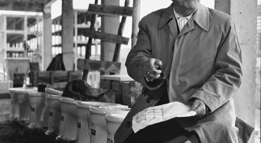 Still from El Verdugo: a man in a long coat looks down at papers in his hands in a half-finished building, in black and white.