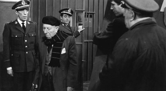 Still from El Verdugo: men in uniform and dark coats stand in an official building, with an elderly man in a beret and scarf at the centre, in black and white.