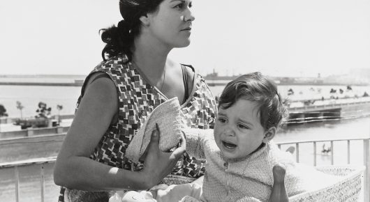 Still from El Verdugo: a black-and-white close-up of a woman in a patterned dress holding a young child, with a seafront behind them.