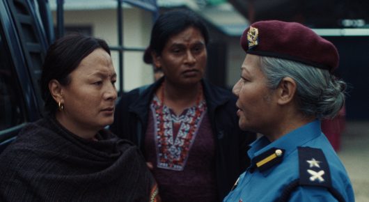 Still from Elephants in the Fog: three women stand close together on a platform, one in a maroon beret and blue uniform, one in a beaded necklace and one in a black shawl.