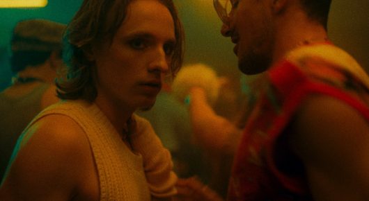 Still from Cambridge Connections: two people stand very close together in a dim, colourful room.