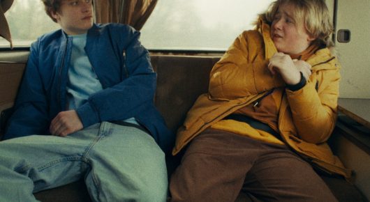 Still from Cambridge Connections: two young people sit side by side on a bench seat in a van, one in a blue jacket and one in an orange-yellow jacket.