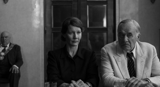 Still from Fatherland: a woman in dark clothes and a man in a pale suit sit side by side in a formal room, with another man seated behind them, in black and white.