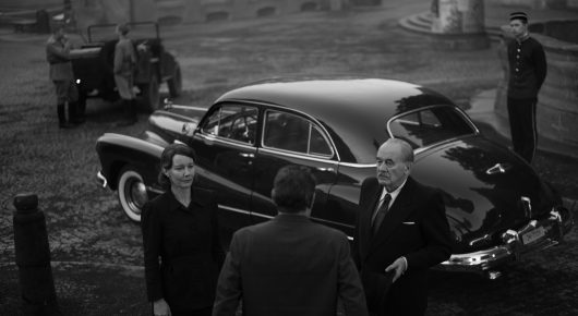Still from Fatherland: a woman and men in dark clothes stand beside a vintage car on a cobbled street, in black and white.