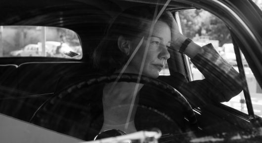 Still from Fatherland: a woman looks out through the window of a vintage car with her hand raised to her head, in black and white.