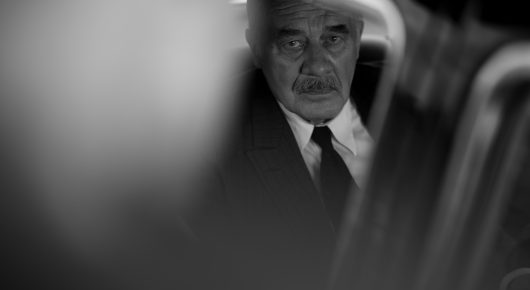 Still from Fatherland: a close-up of an older man with white hair and a dark tie, out of focus except for his face, in black and white.