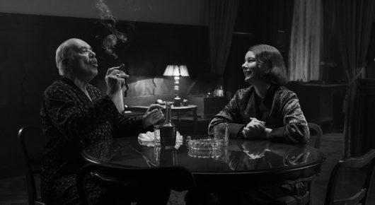 Still from Fatherland: a man smokes a cigarette at a round table opposite a woman in a dim café with a lamp, in black and white.