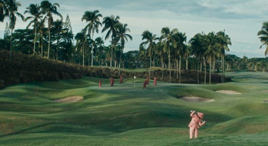 Still from Filipiñana: a wide view of a green golf course with palm trees and a distant figure in pink.