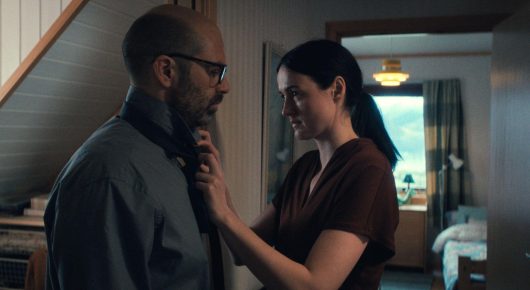 Still from Fjord: a woman reaches up to a man in glasses in a hallway.