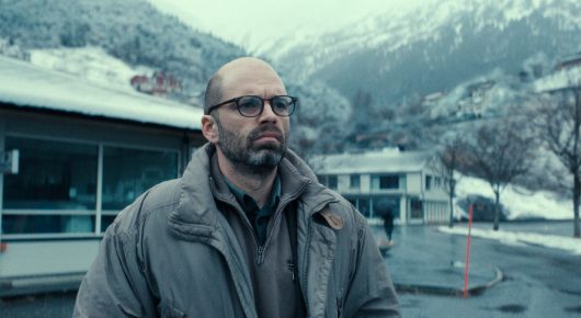 Still from Fjord: a man in a grey jacket stands on a snowy shore in front of a fjord and mountains.