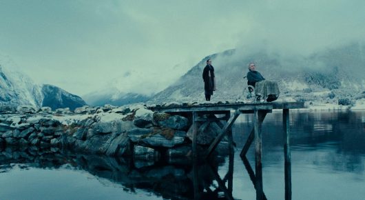 Still from Fjord: two small figures stand on a wooden jetty over still water, with snow-covered mountains behind.